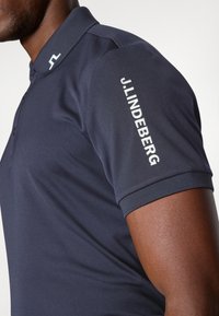 Tmavě modrá polo tričko s klasickým límečkem, krátkými rukávy a nápisem "J.LINDEBERG" v bílé barvě na pravém rukávu. Hladká textura látky.