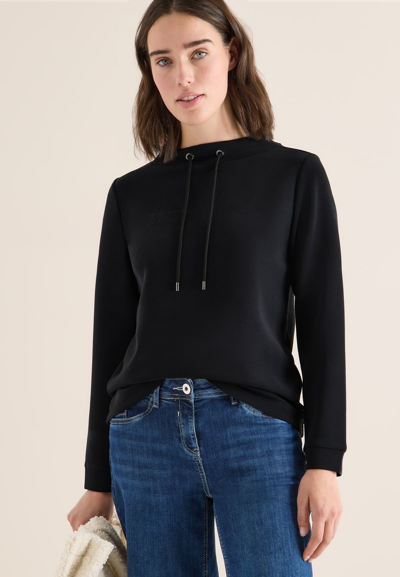 Schwarzer Kapuzenpulli mit Kordelzügen, ausgestattet mit einem strukturierten Logo. Getragen mit blauen Jeans, die einen lässigen, bequemen Sitz zeigen.