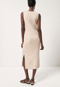 Robe en maille côtelée beige, sans manches, longueur mi-mollet, fente sur le côté et taille cintrée. Texture lisse avec des détails minimalistes.
