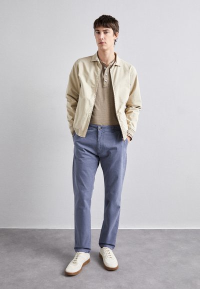 Beige bomberjakke, tan polo-shirt, lyseblå bukser og hvide sneakers med brune såler; afslappet pasform, minimalistisk design og bløde teksturer.