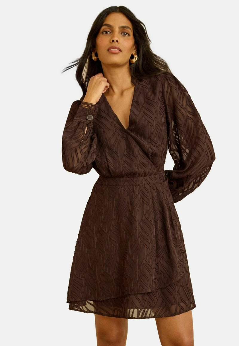 Naine kannab läbipaistvast tumedat pruunist kootud kangast, pikkade varrukatega, v-kaelusega ja kihilise seelikuga kleiti ning poseerib ühe käega näo lähedal.