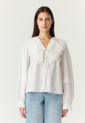 Femme aux cheveux raides et foncés portant un chemisier blanc avec un grand col orné de dentelle et un jean bleu clair sur un fond uni.