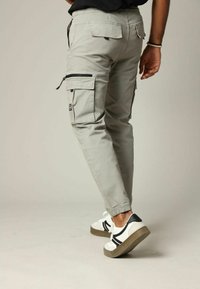 Lichtgrijze cargobroek met elastische boorden, voorzien van zij- en achterzakken, en contrasterende zwarte ritsdetails. G gedragen met witte sneakers.