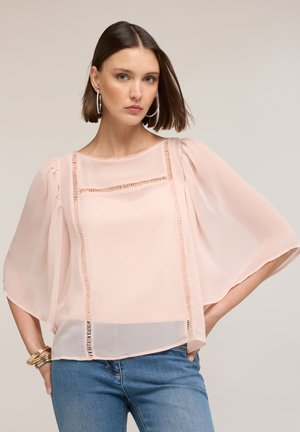 Kobieta o krótkich brązowych włosach, nosząca duże kołowe kolczyki, przezroczystą jasnoróżową bluzkę z szerokimi rękawami oraz niebieskie dżinsy, stojąca z jedną ręką na biodrze.