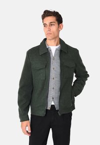 Giacca verde in misto lana con colletto, due tasche sul petto, chiusura frontale con zip e polsini a costine. Indossata sopra una camicia grigia con bottoni.