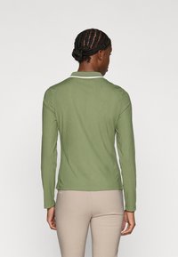 Ilgi piedurkņu polo krekls olīvzaļā ribotas auduma ar baltu apkakles apdari. Iezīmēts ar pieguļošu dizainu un diskrētu logotipu apakšmalā.