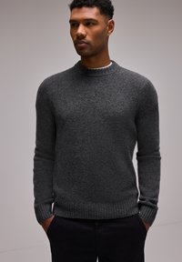 Pull en tricot gris foncé avec un col rond, des poignets et un ourlet côtelés, présentant une finition texturée. Porté avec les mains dans les poches de pantalons noirs.