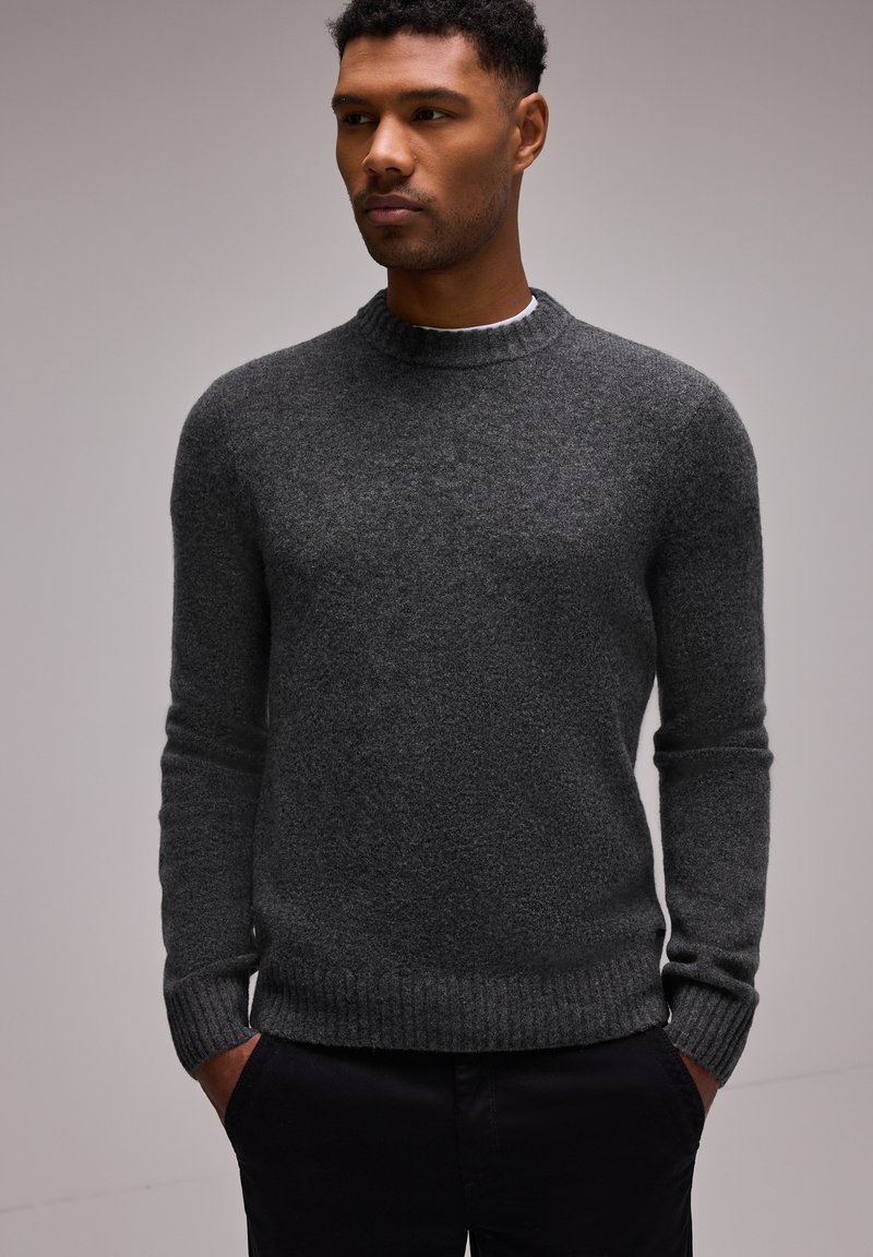 Pull en tricot gris foncé avec un col rond, des poignets et un ourlet côtelés, présentant une finition texturée. Porté avec les mains dans les poches de pantalons noirs.