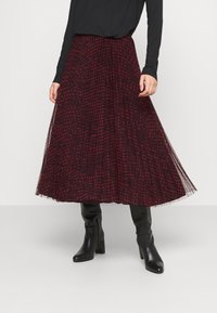 Lang plisseret nederdel i rød og sort ternet mønster, lagdelt med transparent stof. Båret med sorte tætsiddende støvler og en langærmet top.