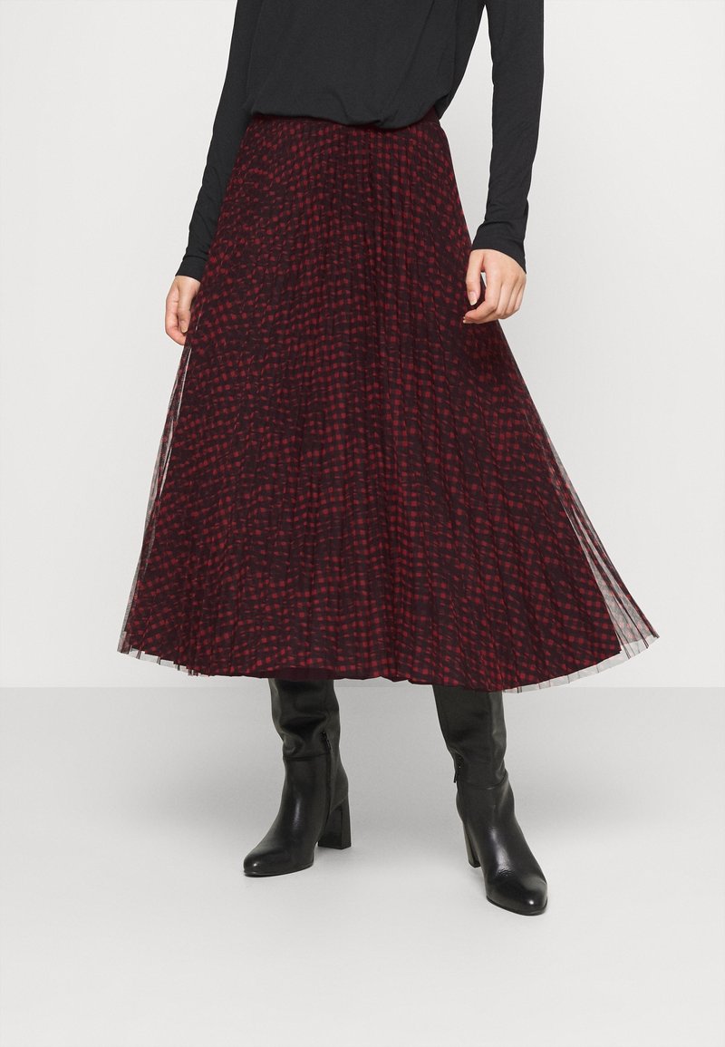 Lang plisseret nederdel i rød og sort ternet mønster, lagdelt med transparent stof. Båret med sorte tætsiddende støvler og en langærmet top.