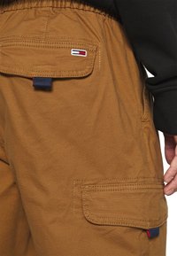 Pantalons cargo marron avec une taille élastique, présentant une poche arrière et une étiquette de logo en rouge, blanc et bleu. Texture lisse avec des détails de couture.