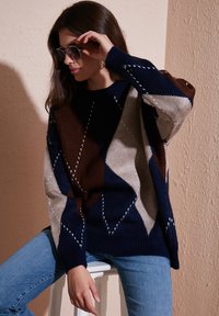 Knitted sweater with a geometric diamond pattern in navy, brown, and beige. Ribbed cuffs and hem, paired with light blue jeans.