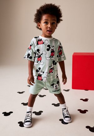 Lille barn står på gulvet med Mickey Mouse-hovedmønstre, iført matchende Mickey Mouse-t-shirt og shorts samt sorte sneakers.