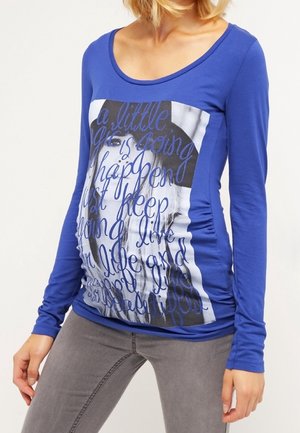 Kobieta ubrana w niebieską koszulę z długim rękawem z czarno-białym zdjęciem i nadrukiem w stylu pisma kursywą, w połączeniu z szarymi spodniami, ręce swobodnie opuszczone wzdłuż ciała.