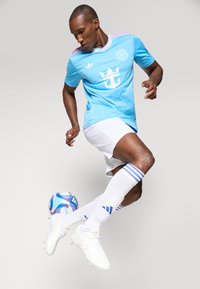 Camisola de futebol azul claro com detalhes rosa, calções brancos e chuteiras brancas. Jogador conduz uma bola com padrão de desenhos em azul e vermelho.