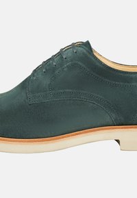 Scarpa in suede verde scuro con una texture liscia, caratterizzata da un design con lacci, dettagli cuciti e una suola di colore chiaro.