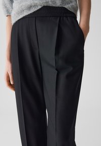 Pantalons noirs en tissu texturé, avec un devant plissé et une taille élastique. Des poches latérales sont visibles, améliorant la fonctionnalité.