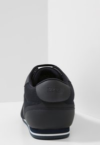 Marinblå sneaker med meshuvud, gummihäl och märkeslogotyp. Har en texturerad yta och en kontrasterande vit rand på sulan.