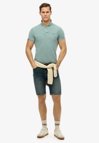 Polo shirt azzurro chiaro a maniche corte, pantaloni denim, maglione crema legato in vita, sneakers beige e calzini a fantasia.