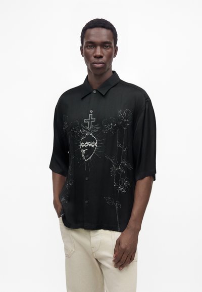 Homme portant une chemise noire à boutons avec un motif Sacré-Cœur blanc et floral, associée à un pantalon beige clair, sur fond blanc.