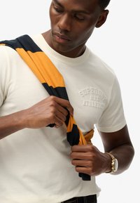 Camiseta blanca de algodón con logo de la marca en relieve, con un suéter a rayas azul marino y naranja caído sobre el hombro. Reloj marrón en la muñeca.