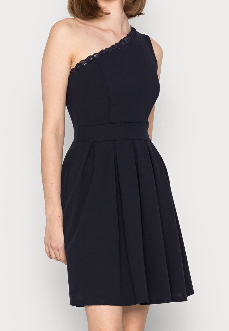 Robe navy à une épaule avec bordure en dentelle texturée, corsage ajusté, jupe plissée et coupe à hauteur des genoux. Tissu lisse avec un éclat subtil.
