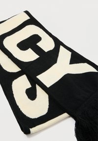 Sciarpa nera e crema con grandi lettere a blocchi e bordo frangiato. Realizzata in tessuto morbido con colori a contrasto e texture liscia.