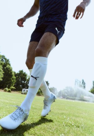 Fehér foci cipők szürke részletekkel, tengerészkék sportnadrág és fehér, térd fölé érő zoknik logóval, egy fűves pálya háttérben.