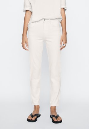White slim-fit jeans with a button closure, five pockets, and subtle stitching. Paired with a loose, light top and black flip-flops.