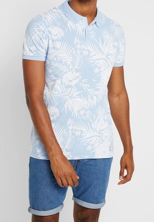 Man wearing light blue polo shirt with white tropical floral print and cuffed denim shorts, standing with arms relaxed.