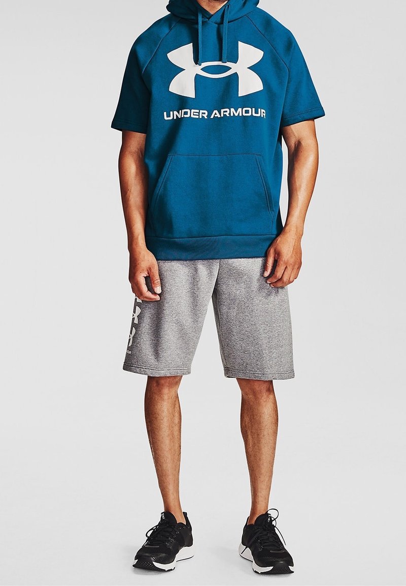 Tyrkysová mikina s kapucí a velkým bílým logem Under Armour, v kombinaci se šedými kraťasy a černými sportovními botami. Tkanina vypadá hladce a jemně.