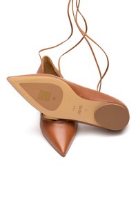 Scarpe in pelle marrone a punta con sottili cinturini alla caviglia. Texture liscia con una suola leggera caratterizzata da dettagli dorati di branding e logo.