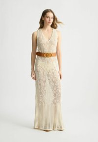 Abito in crochet crema, lunghezza ginocchio, con scollatura a V e motivo a diamante, abbinato a una cintura larga color tan con una grande fibbia.