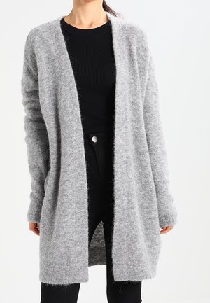 Kvinde iført en lang, åben, lysegrå, blød cardigan over en sort ribbet top og sorte bukser mod en ensartet baggrund.