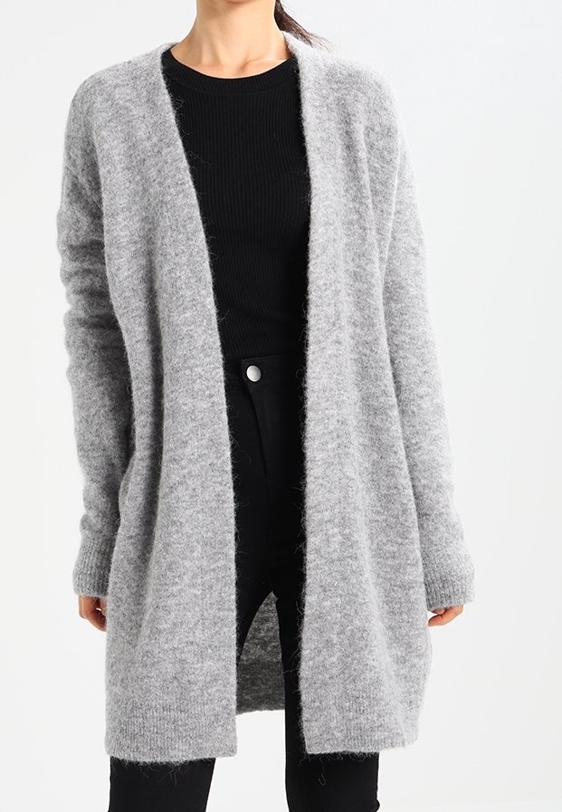 Femme portant un long cardigan gris clair en matière pelucheuse, ouvert sur le devant, par-dessus un haut noir côtelé et un pantalon noir, sur un fond uni.