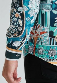 Manche et poignet d'une chemise à motifs avec des designs floraux et architecturaux en teal, noir, orange et blanc, portée avec un pantalon noir.