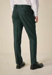 Green textured trousers with two back pockets and a flat front. Paired with black dress shoes, the fabric appears slightly woven.