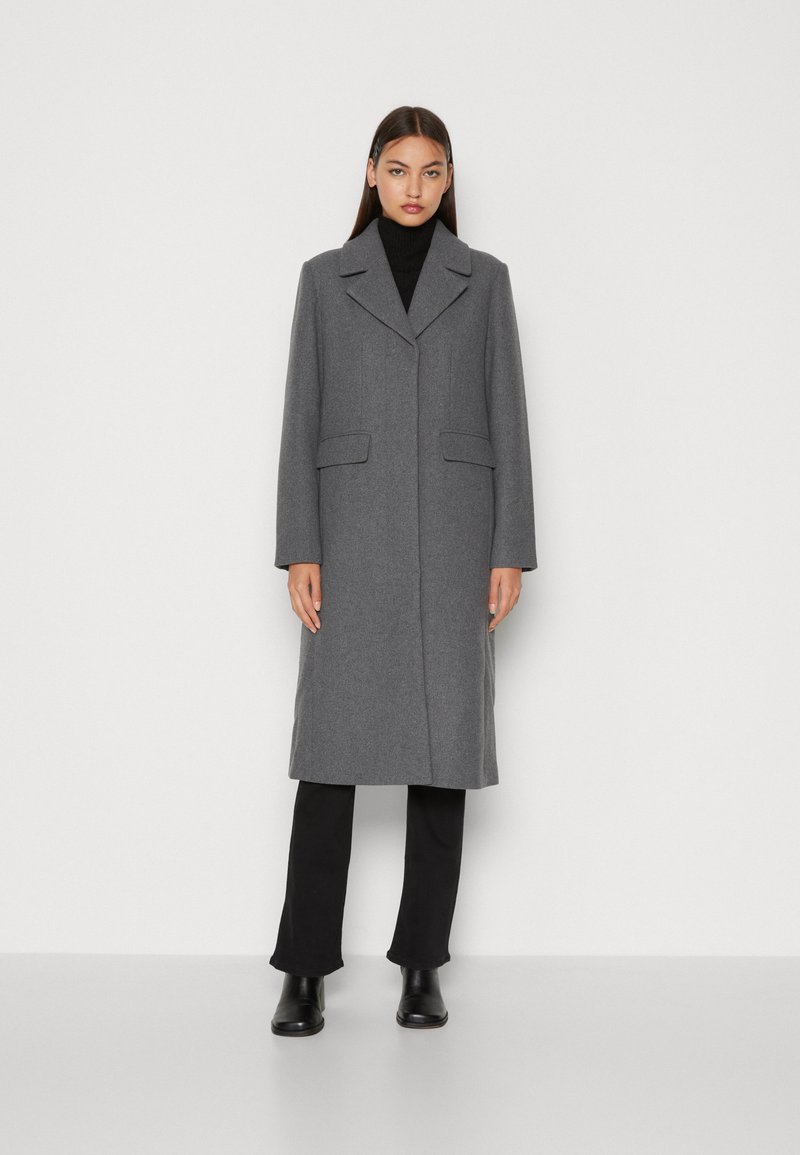 Manteau en laine gris avec revers crantés, deux poches avant et une silhouette droite. Porté sur un col roulé noir et un pantalon.