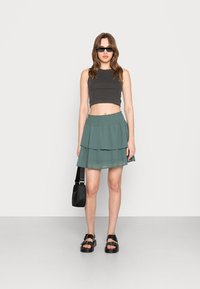 Top cropped cinza canelado, saia teal em camadas, sandálias pretas com fivela, pequena bolsa preta à tiracolo, fundo plain.