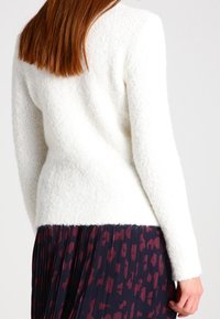 Femme aux cheveux roux lisses portant un pull blanc texturé à manches longues et une jupe plissée sombre avec des motifs abstraits rouges.