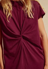 Haut bordeaux à manches courtes avec un nœud torsadé au centre, fabriqué dans un tissu doux, offrant une coupe décontractée et une texture lisse.