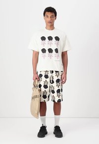 Hvit t-skjorte med svart blomstermønster, sammen med kremfargede shorts med svart og grønt blomstermønster. Modellen holder en beige veske.