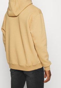 Persona vestida con una sudadera con capucha beige y jeans negros, vista desde atrás sobre un fondo claro y liso.