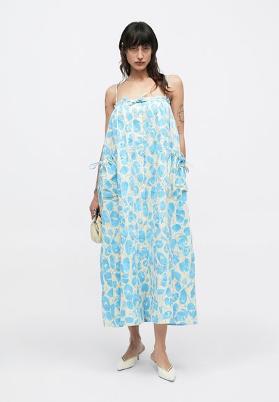 Μακρύ φόρεμα ανοιχτό μπλε με λουλουδάτα σχέδια, κατασκευασμένο από αέρινο ύφασμα. Διαθέτει ρυθμιζόμενους ιμάντες και δεσίματα στα πλαϊνά. Φορεμένο με μια μικρή τσάντα.