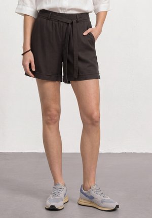 Ben iført sorte shorts med snører, hvid skjorte stoppet ind og grå sneakers, stående på en lysegrå gulv mod en hvid væg.