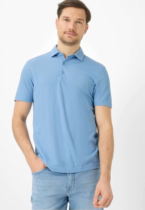 Mand iført lyseblå kortærmet poloshirt og lyseblå jeans, stående med den ene hånd i lommen mod en hvid baggrund.