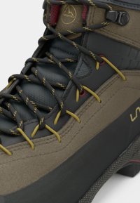 Brūna un pelēka pārgājienu zābaku ar tekstūrētu augšdaļu, dzeltenām un melnām auklām, kā arī izturīgu zoli ar sarkanajiem akcentiem, kas nodrošina saķeri.