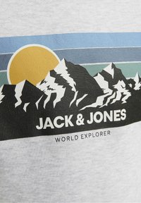 Šedé tričko s grafickým motivem hory v černé, bílé a zelené barvě, se žlutým sluncem a modrými pruhy, označené "JACK & JONES WORLD EXPLORER."
