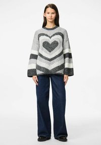 Maglione grigio e bianco con motivo a cuore, dotato di maniche ampie e una texture morbida, abbinato a jeans a gamba larga blu scuro.