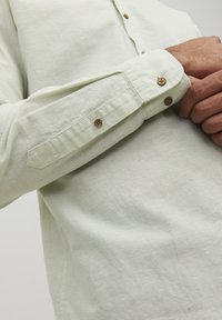 Camisa de botones de color verde claro con una textura suave, mangas largas con puños abotonados y un corte relajado.